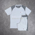 A white and gray athletic short-sleeve shirt and matching shorts laid flat on a gray concrete surface. The shirt has gray shoulder panels and a small chest logo, while the shorts have gray side panels and an elastic waistband.
